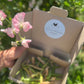 Cardboard box with a collection of sweet pean pods, in a box with a label, held in front of pink flowers and greenery.